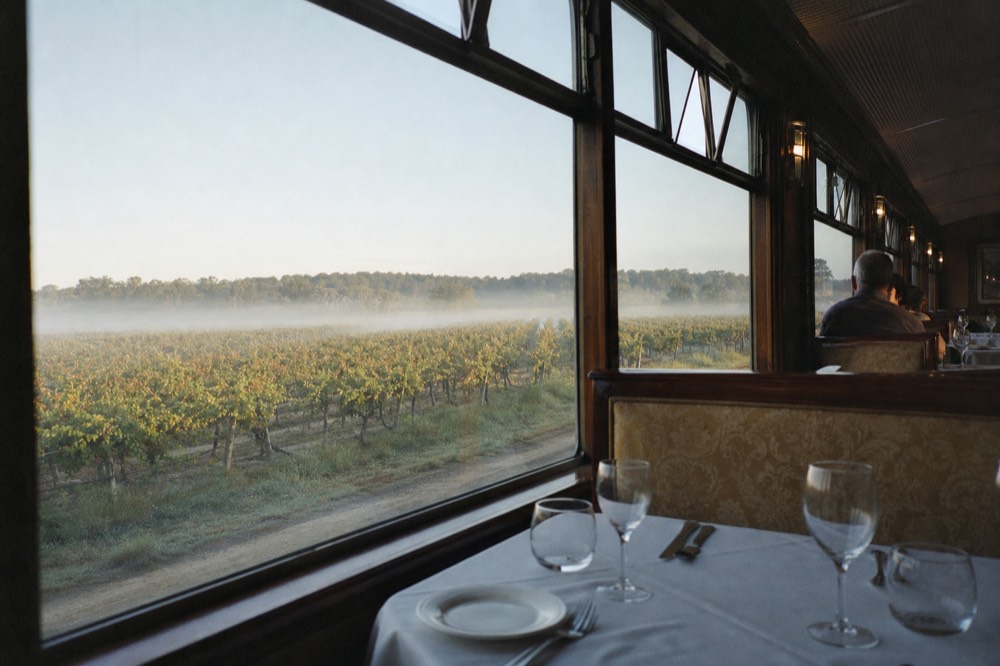 Interior of a vintage dining car with white tablecloths and glassware beside large windows framing rows of vines, no text, no logos, no faces