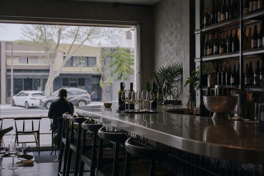 A downtown storefront tasting room with a polished bar, stools and bottles on a back shelf, bright street light through the window, no text, no logos, no faces