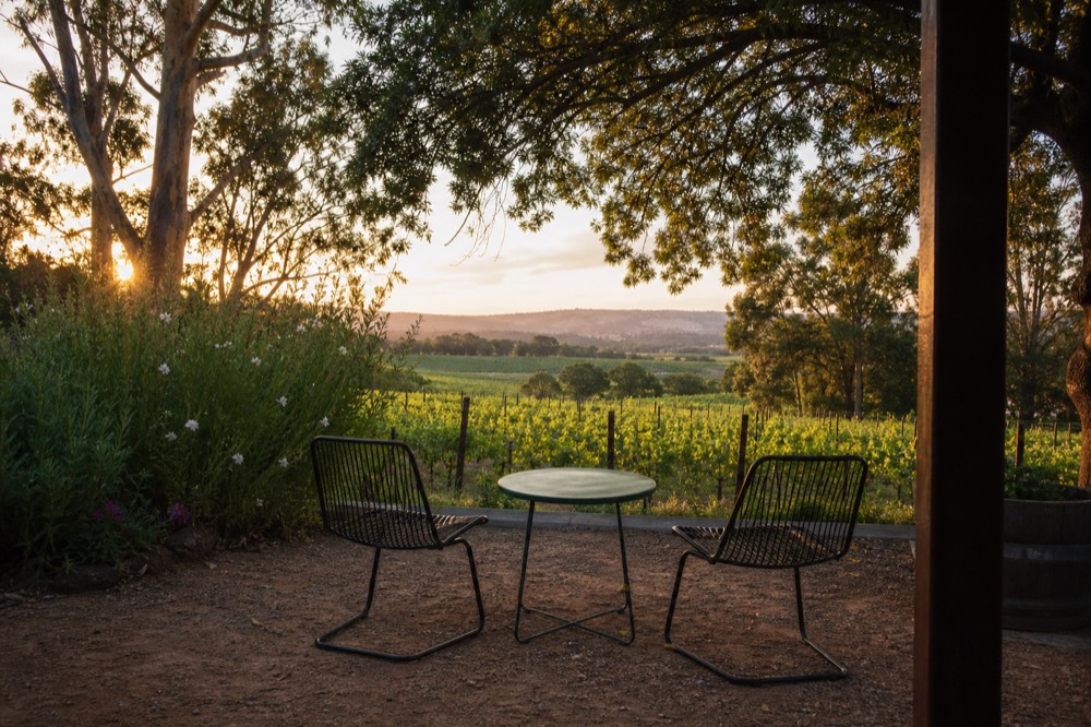A couple's-eye view of a quiet vineyard terrace with two empty chairs facing rows of vines and distant hills at sunset, no text, no logos, no people